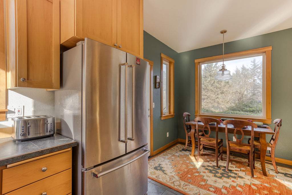 Kitchen area with a stainless steel refrigerator, light wood cabinetry, and a small breakfast nook with a wooden table and chairs by a window, all set on a tiled floor.