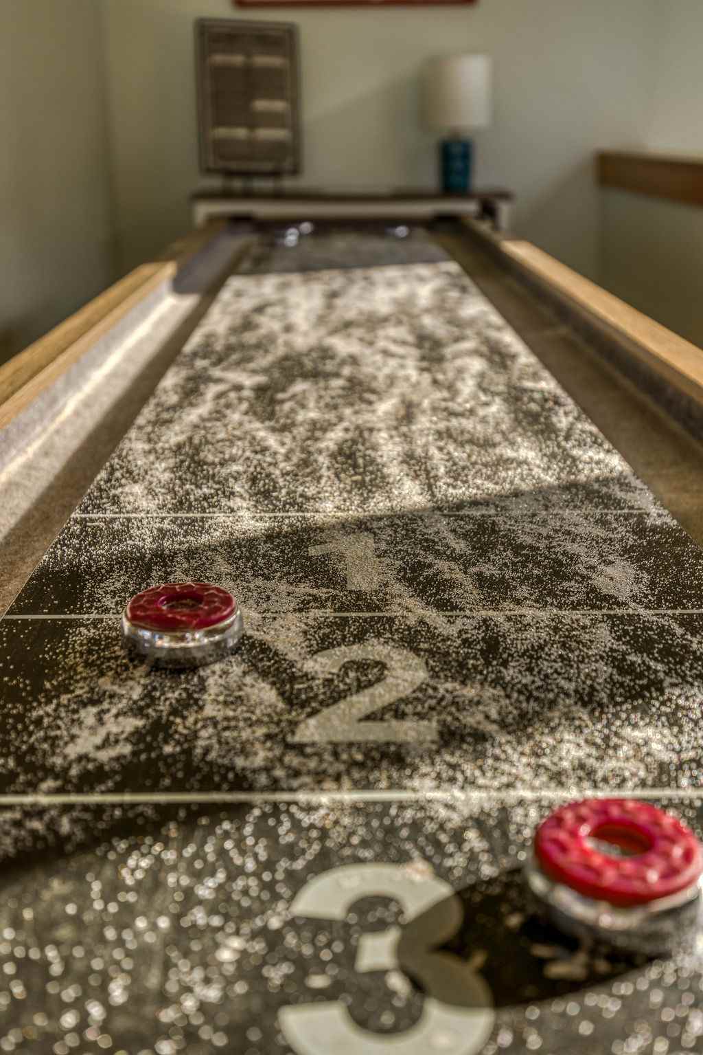 Close-up view of a shuffleboard table with red pucks, showing the textured playing surface with score markers. The room appears to be a dedicated game area.