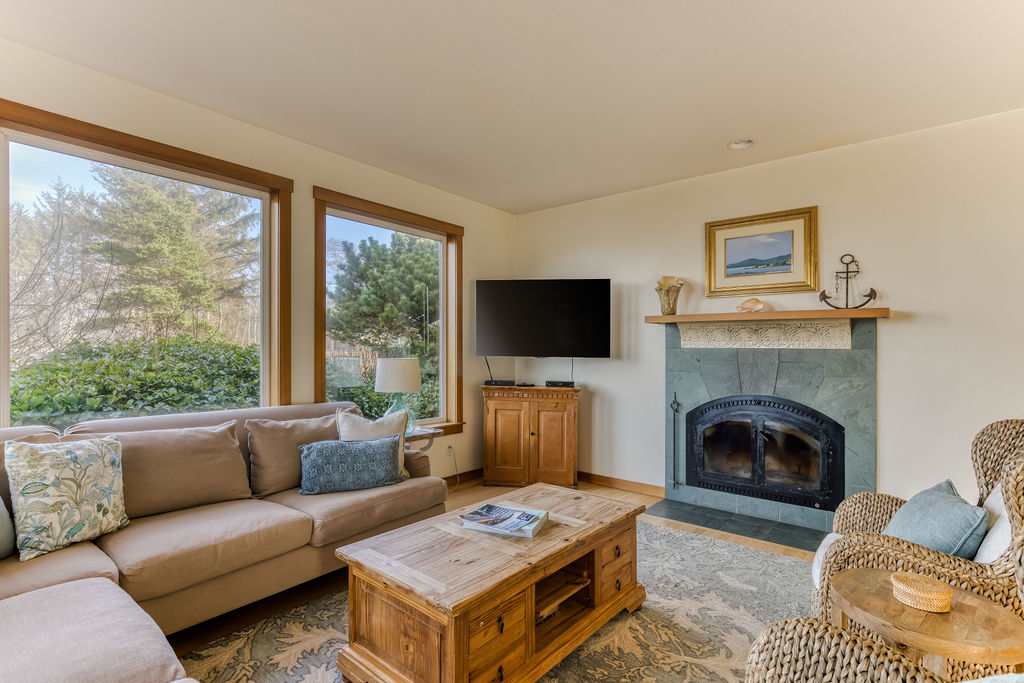 Inviting living room with a beige sectional sofa and a wicker armchair facing a stone-tiled fireplace with a mounted flat-screen TV, all set on a patterned rug with large windows overlooking greenery.