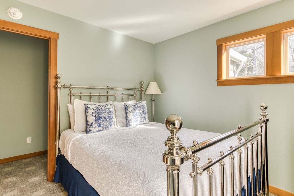 A bedroom with light green walls, a polished metal bed frame with white bedding and blue patterned pillows, and small windows with natural wood trim.