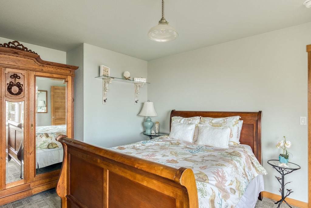 A cozy bedroom featuring a wooden sleigh bed with white bedding, light blue walls, a large wooden armoire with a mirror, and a small glass-top side table with a lamp.