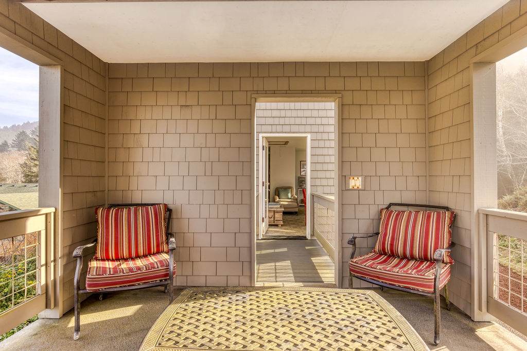 A cozy covered balcony within the lookout tower, featuring two striped outdoor armchairs and a small metal table. The walls are clad in light brown shingles, and a doorway leads back into the main house.