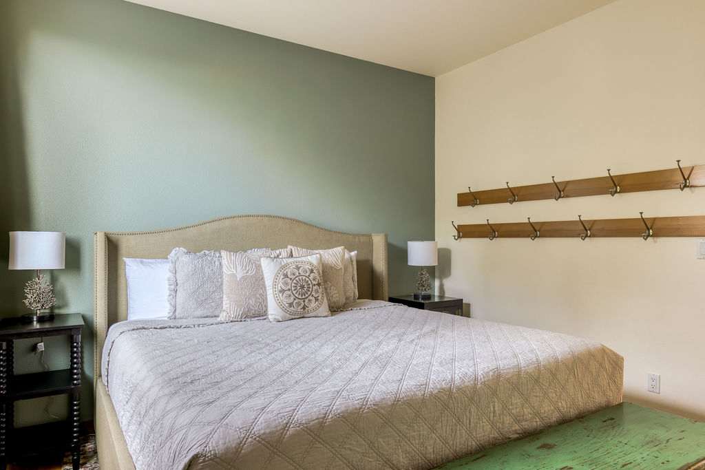 This bedroom features a large bed with a beige upholstered headboard and light-colored quilted bedding. The walls are painted in a two-tone scheme of light green and cream, complemented by wooden coat racks.