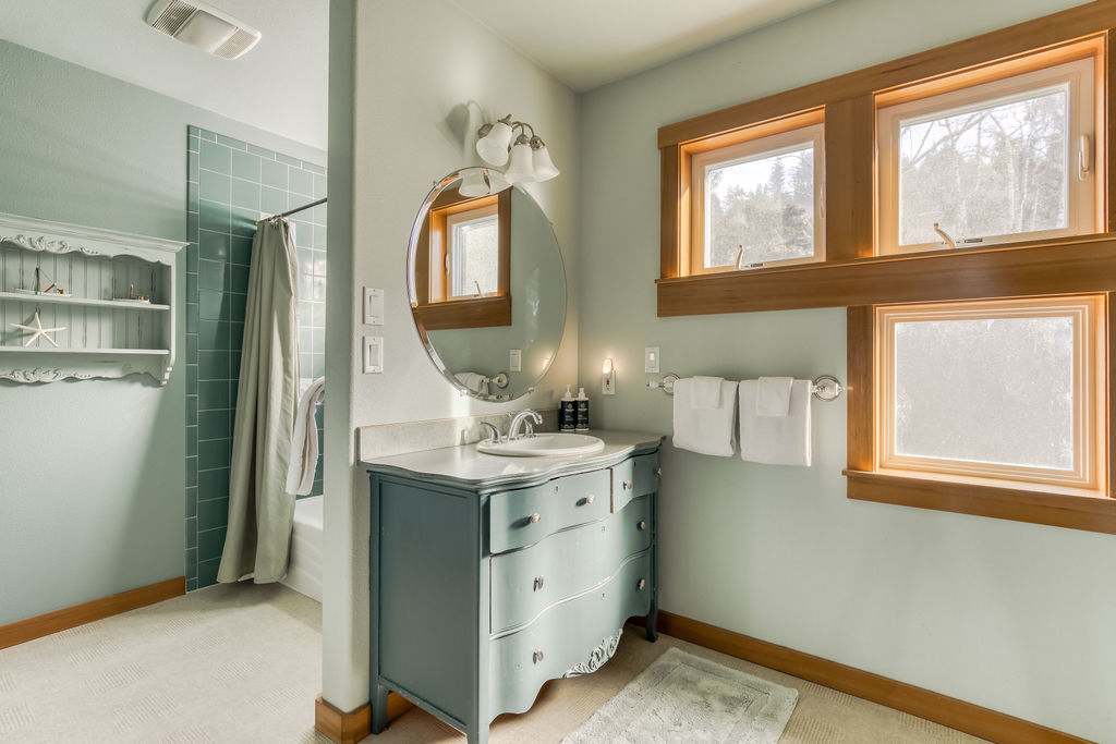 A bathroom with light blue walls, a unique light blue dresser vanity with a round mirror, and a shower/tub combo visible behind a partial wall with green tiled walls.