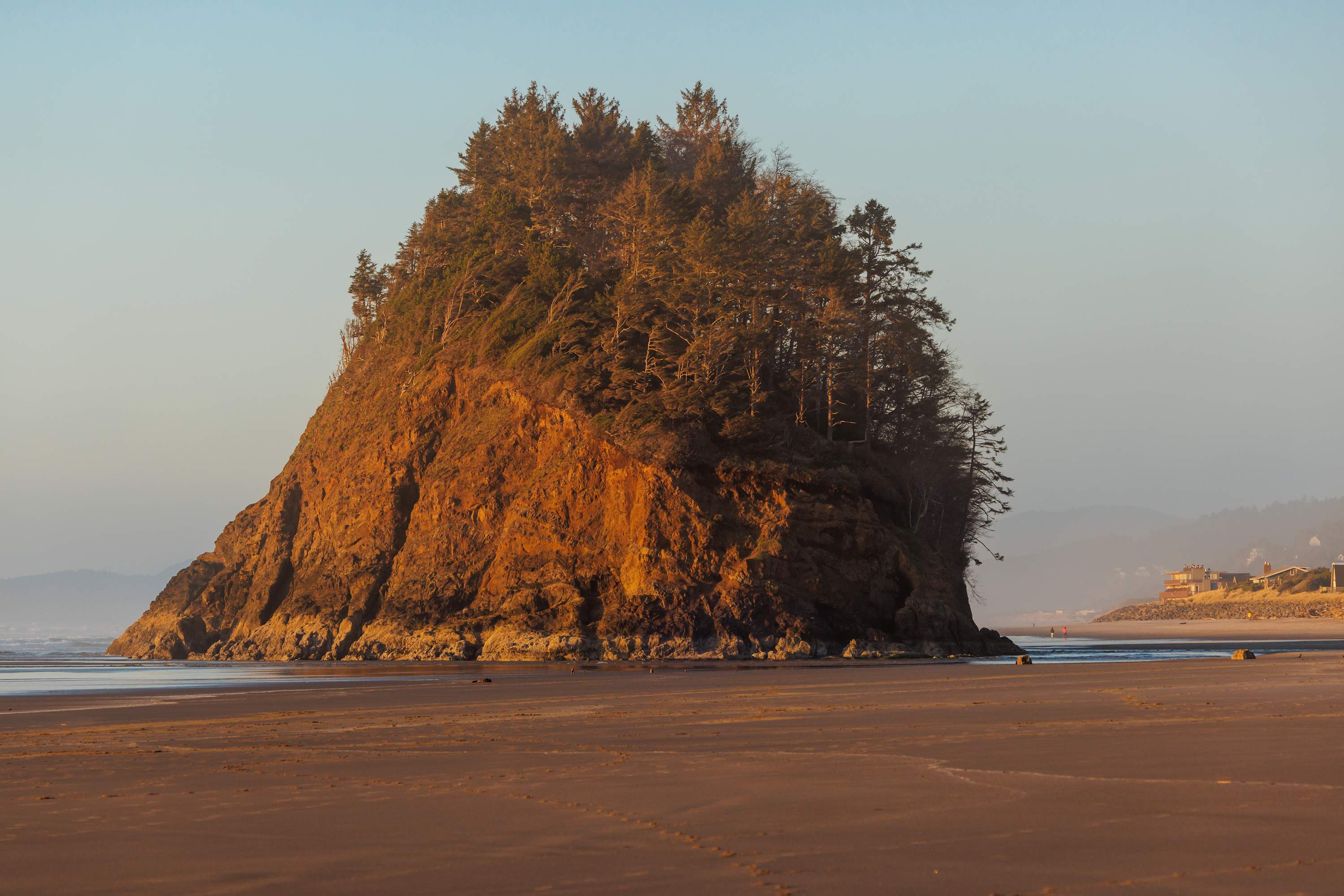 A vast sandy beach at sunset, dominated by a large, tree-covered sea stack (Proposal Rock) in the midground, with distant houses visible along the coastline under a clear sky.