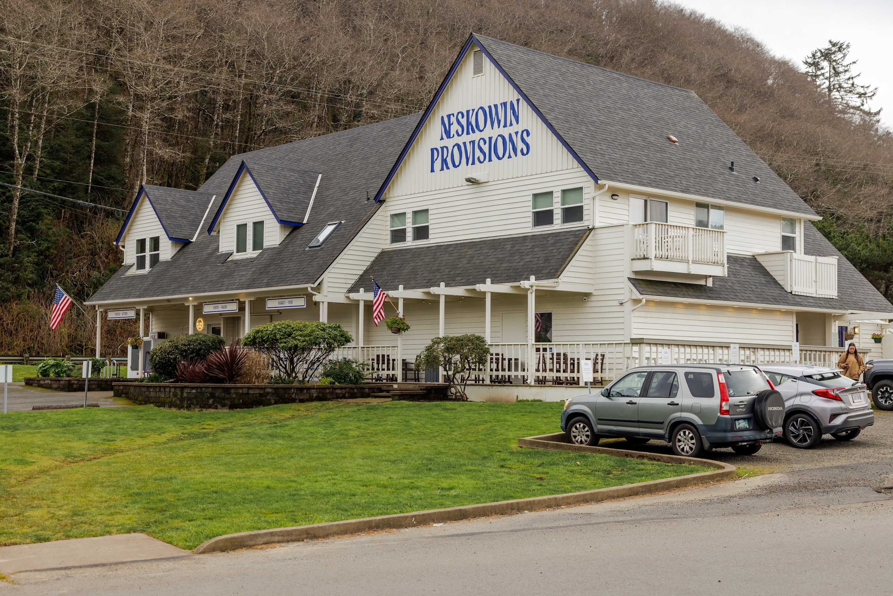 A large white building with blue trim and a dark roof, featuring "NESKOWIN PROVISIONS" signage, a covered porch with outdoor seating, and cars parked in front on a grassy lawn.