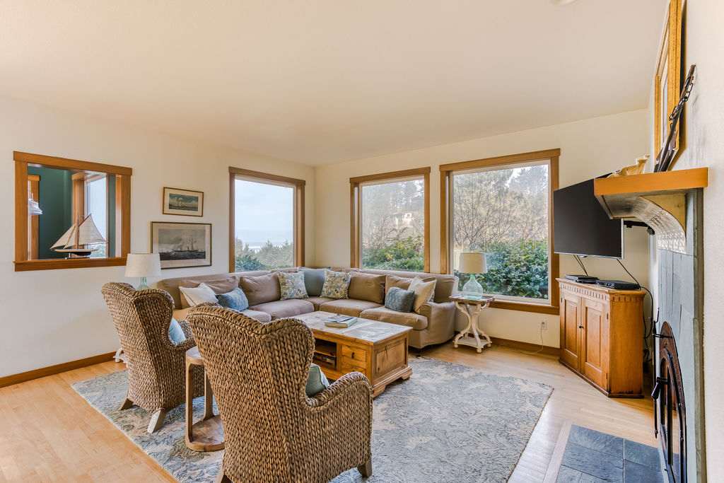Cozy living room featuring a large L-shaped sectional sofa with throw pillows, two wicker armchairs, and a rustic wooden coffee table on a patterned rug, with multiple windows offering outdoor views. A flat-screen TV is mounted near a fireplace.