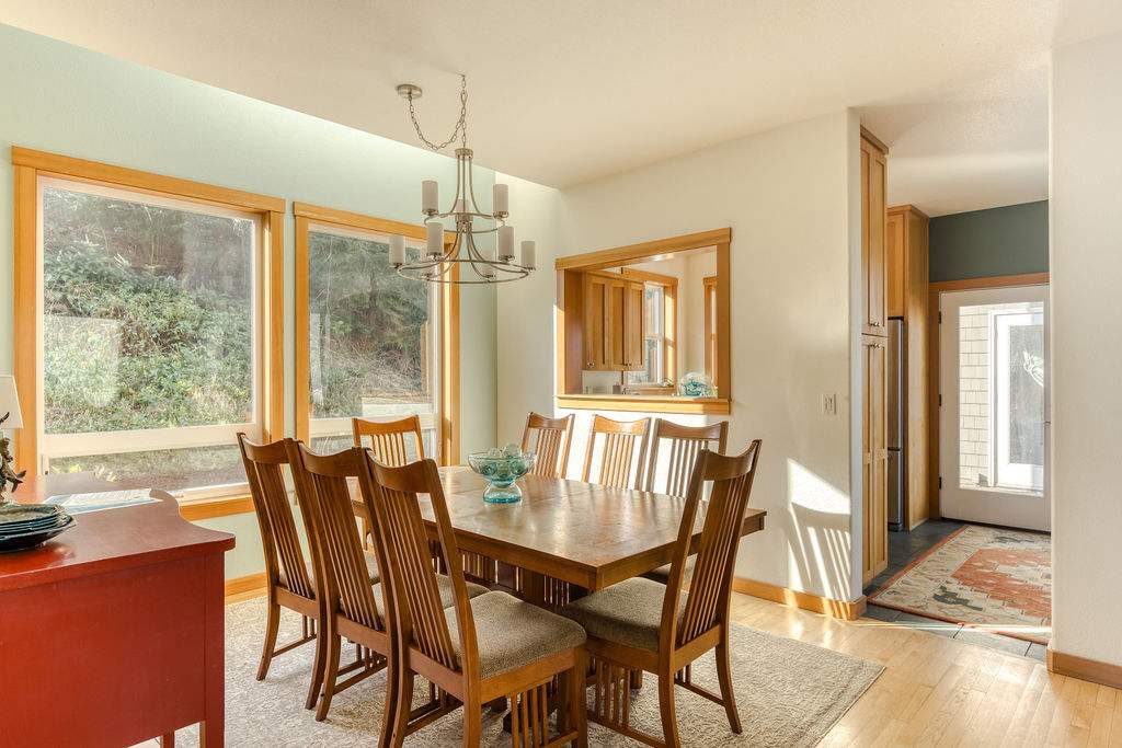 Bright dining room with a wooden table and chairs, illuminated by natural light from large windows, with a pass-through window to the kitchen and a view of the entryway.