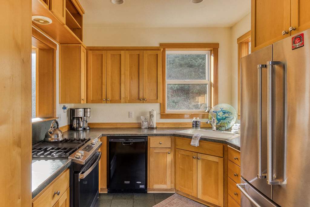 Well-equipped kitchen featuring light wood cabinets, dark countertops, a gas range, dishwasher, and a stainless steel refrigerator, with a window above the sink offering a glimpse of the outdoors.