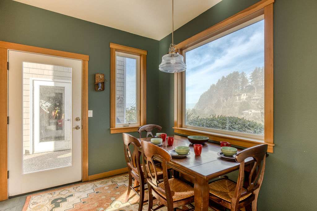 A cozy dining area features dark green walls, a wooden table set for four, and large windows offering a stunning ocean view of a tree-covered hill. A clear glass pendant light hangs above the table.