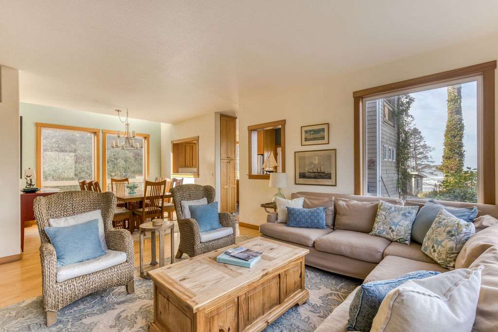 Bright living room featuring a large L-shaped sectional sofa, two wicker armchairs, and a rustic wooden coffee table on a patterned rug, with a dining area visible in the background. Large windows offer views of the outdoors.