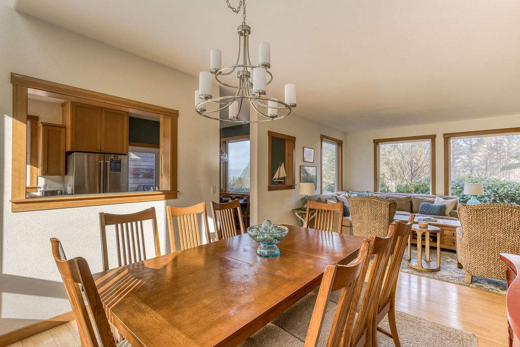 Elegant dining room featuring a long wooden table with eight chairs and a modern chandelier, with a pass-through window to the kitchen and the living room visible in the background.