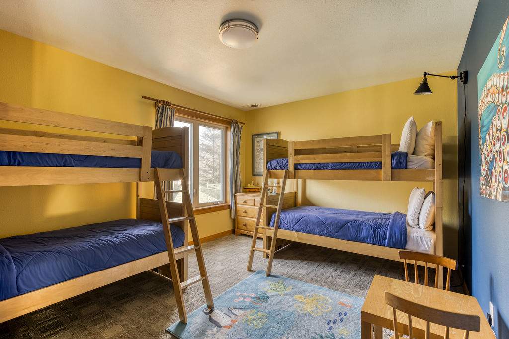 A vibrant bedroom with yellow walls, two wooden bunk beds with blue bedding, a patterned rug on the carpeted floor, and a window with curtains.
