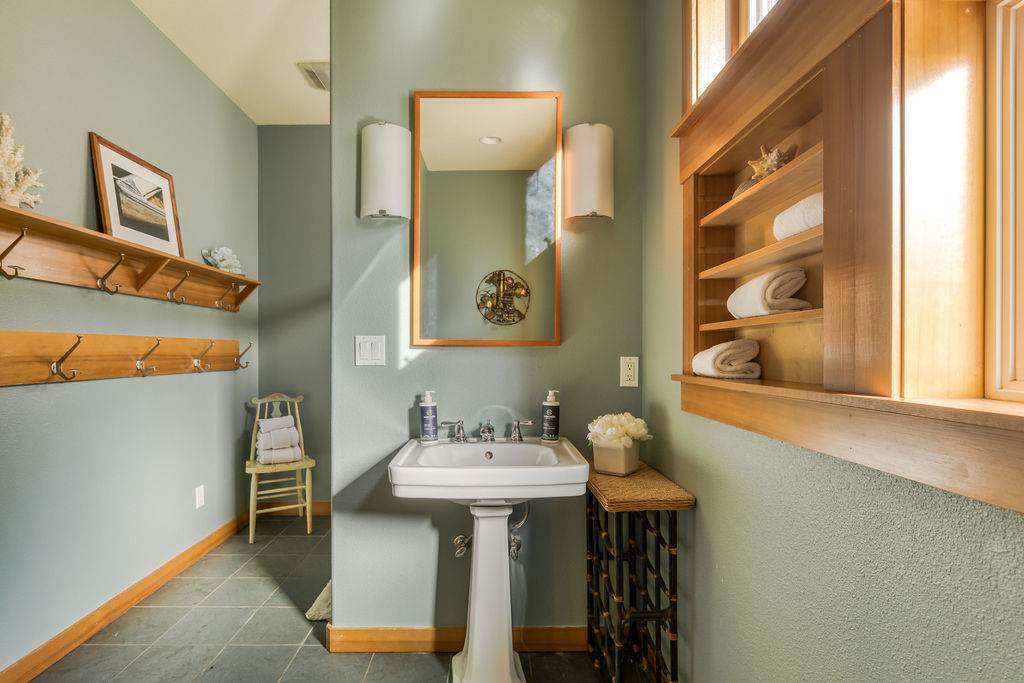 The bathroom features light green walls, a classic pedestal sink, and a large mirror with a warm wooden frame. A built-in wooden shelf unit neatly holds towels, and a small window provides natural light.