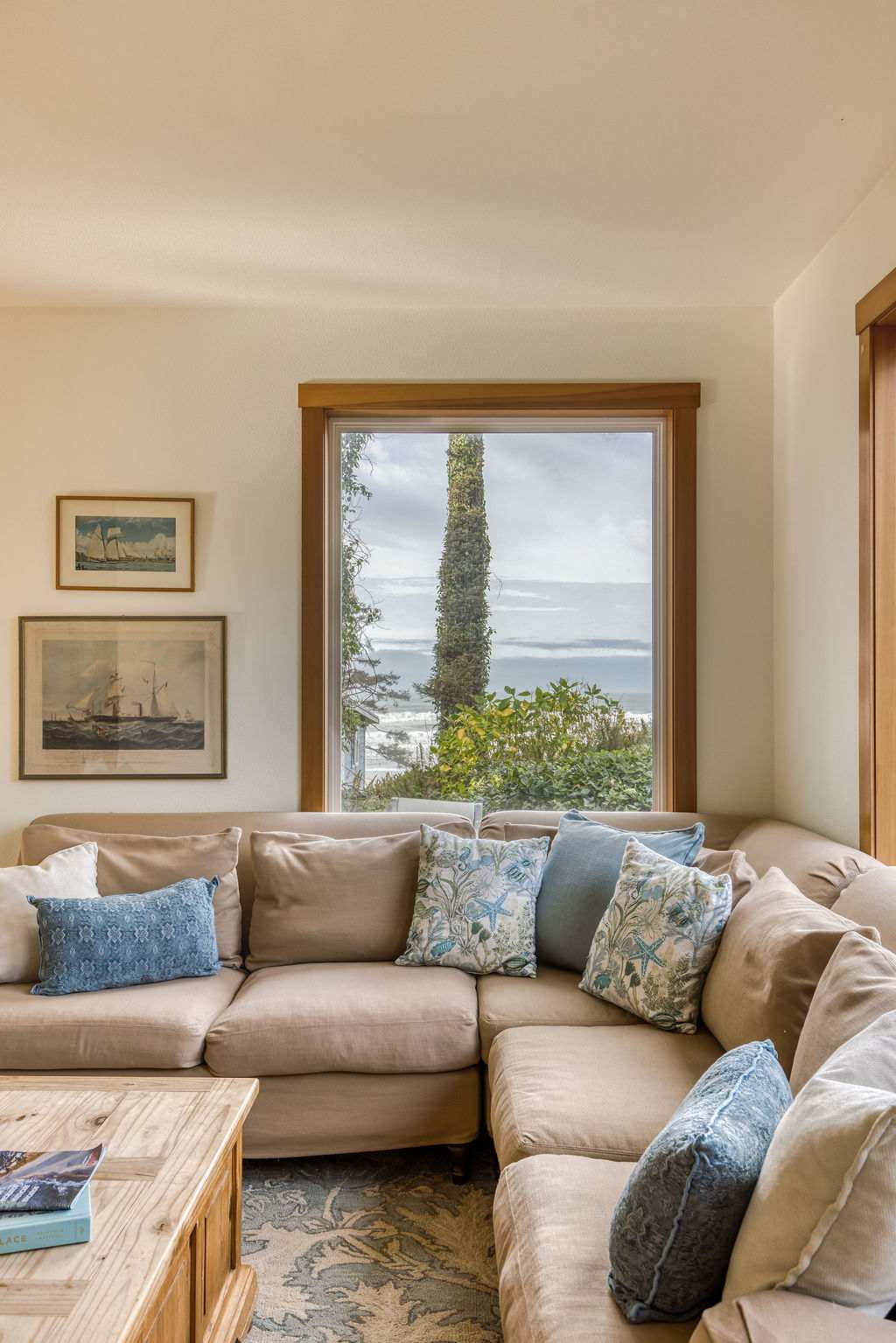 Cozy living room featuring a large L-shaped beige sectional sofa with blue and patterned throw pillows, facing a window with a partial ocean view. Two framed nautical prints adorn the light-colored wall.