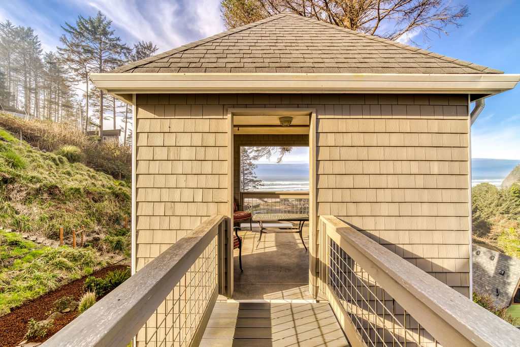 A covered wooden walkway with railings, leading to a lookout tower with an opening that frames a stunning ocean view in the distance. The structure is clad in light brown shingles, surrounded by green hills and trees under a bright sky.