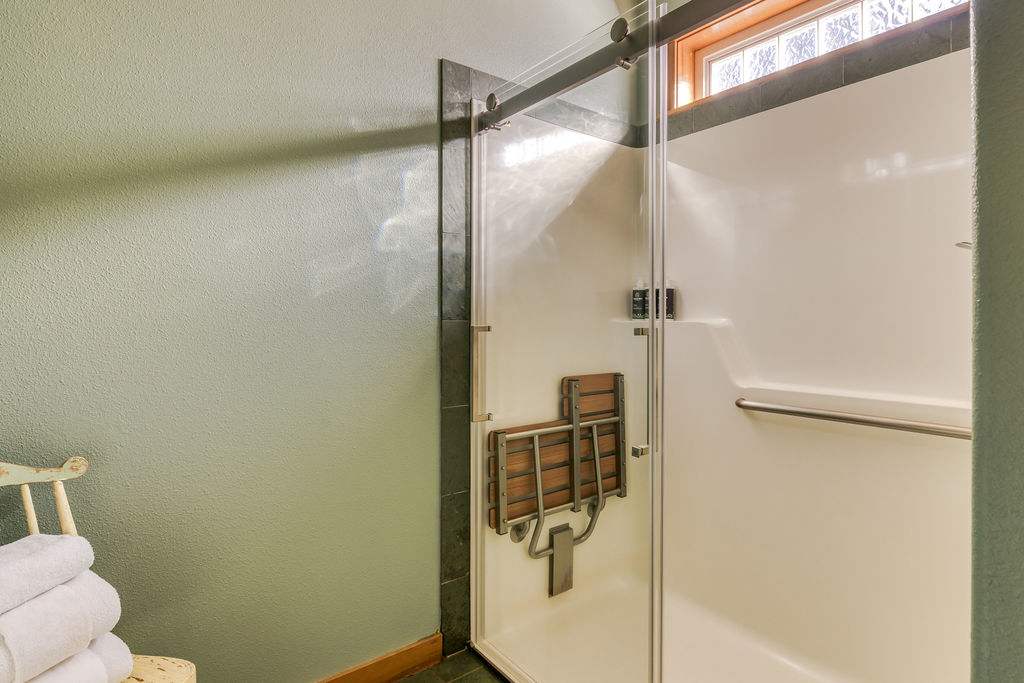 A modern walk-in shower features a glass sliding door, dark gray tile accents, and a convenient built-in wooden fold-down seat. The shower has a clean white surround and a small window above for natural light.