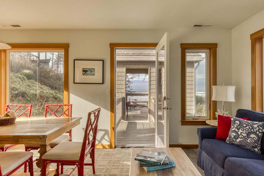 A bright dining area with a light wood table and red chairs, adjacent to a living space with a blue sofa. A glass door opens to an outdoor deck, revealing a distant ocean view and lush greenery outside the large windows.