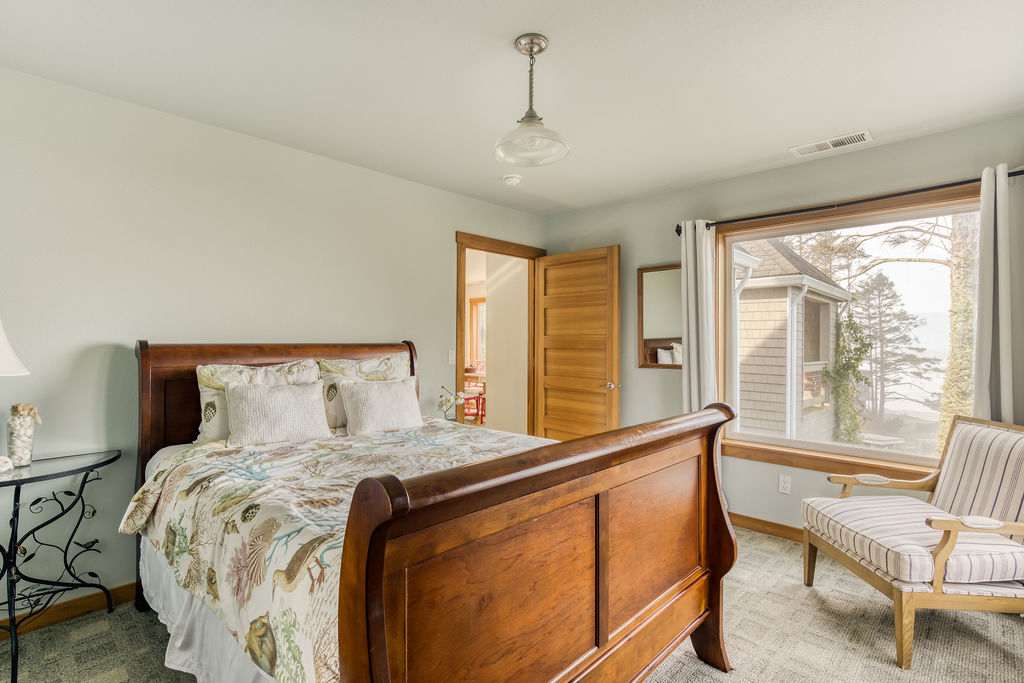 A bright bedroom featuring a wooden sleigh bed with a patterned duvet, light blue walls, and a large window offering a partial view of the outdoors and another building.