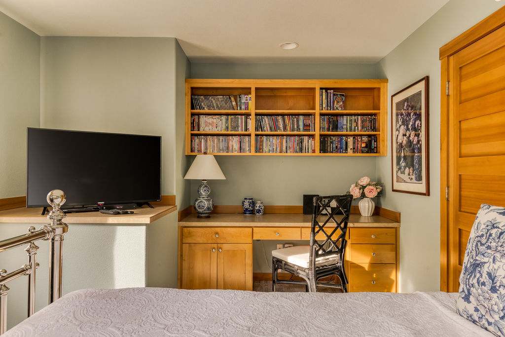 A desk area in bedroom-3 featuring a built-in wooden desk with shelves filled with books and DVDs, a black chair, and a flat-screen TV on a shelf.