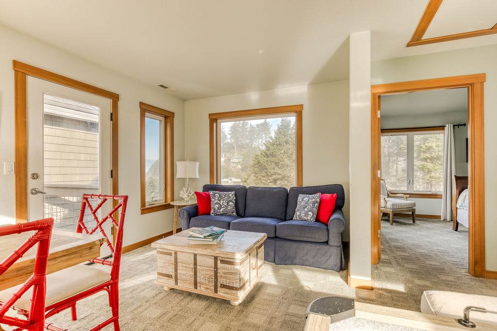 A comfortable living room featuring a blue sofa with red and patterned throw pillows, and a rustic wooden coffee table. Large windows and a glass door offer views of the outdoors, and a doorway leads to an adjacent bedroom.