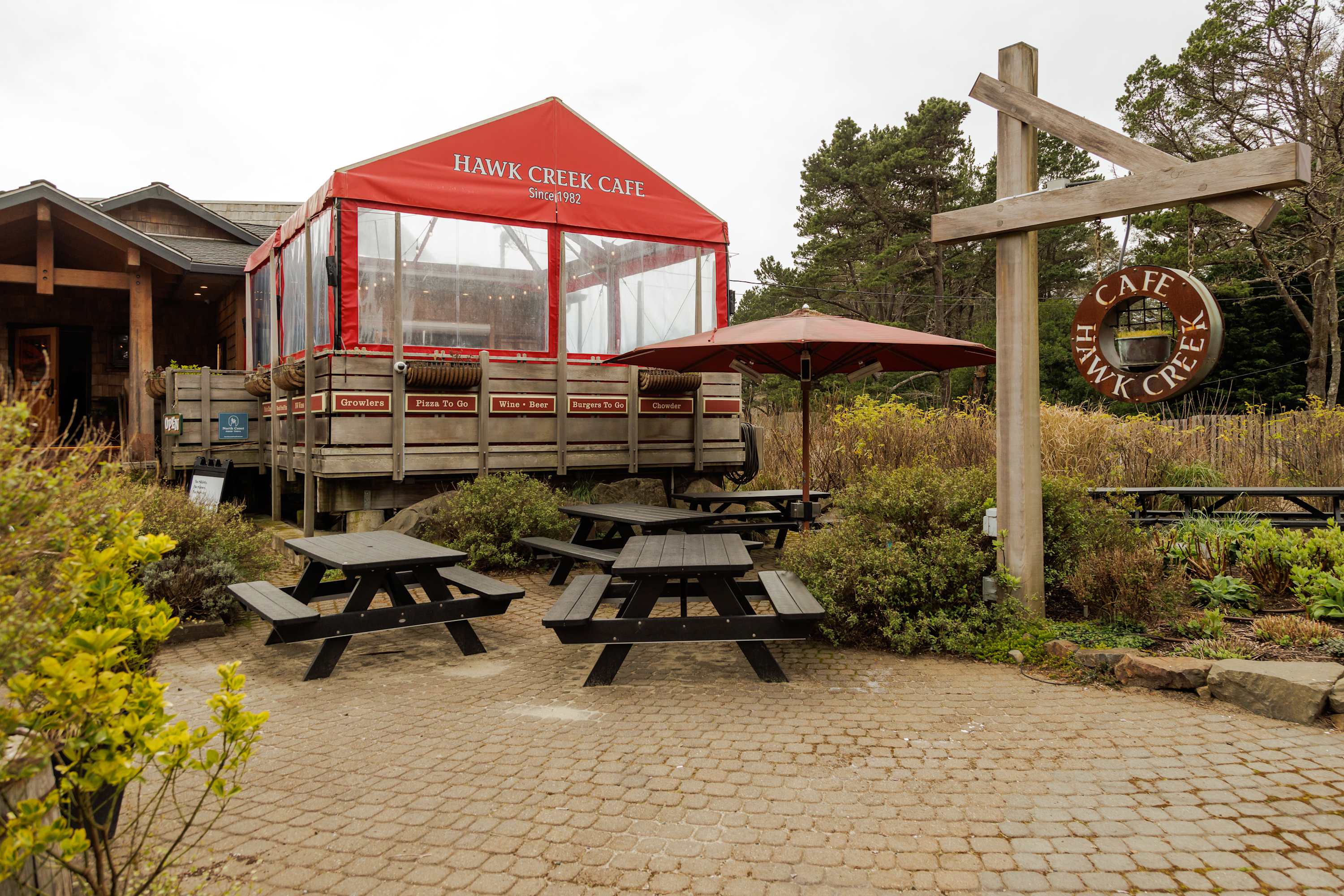 An outdoor patio area with black picnic tables on a cobblestone surface, adjacent to a building with a red awning labeled "HAWK CREEK CAFE" and surrounded by lush greenery.