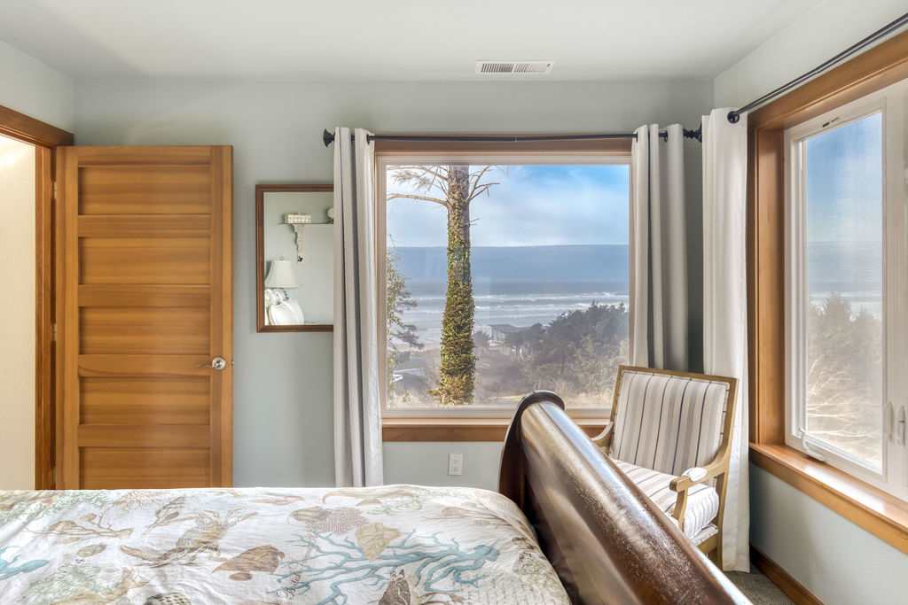 A close-up view of the large window in bedroom-1, showcasing a stunning ocean view with a sandy beach and trees, framed by white curtains and natural wood trim.