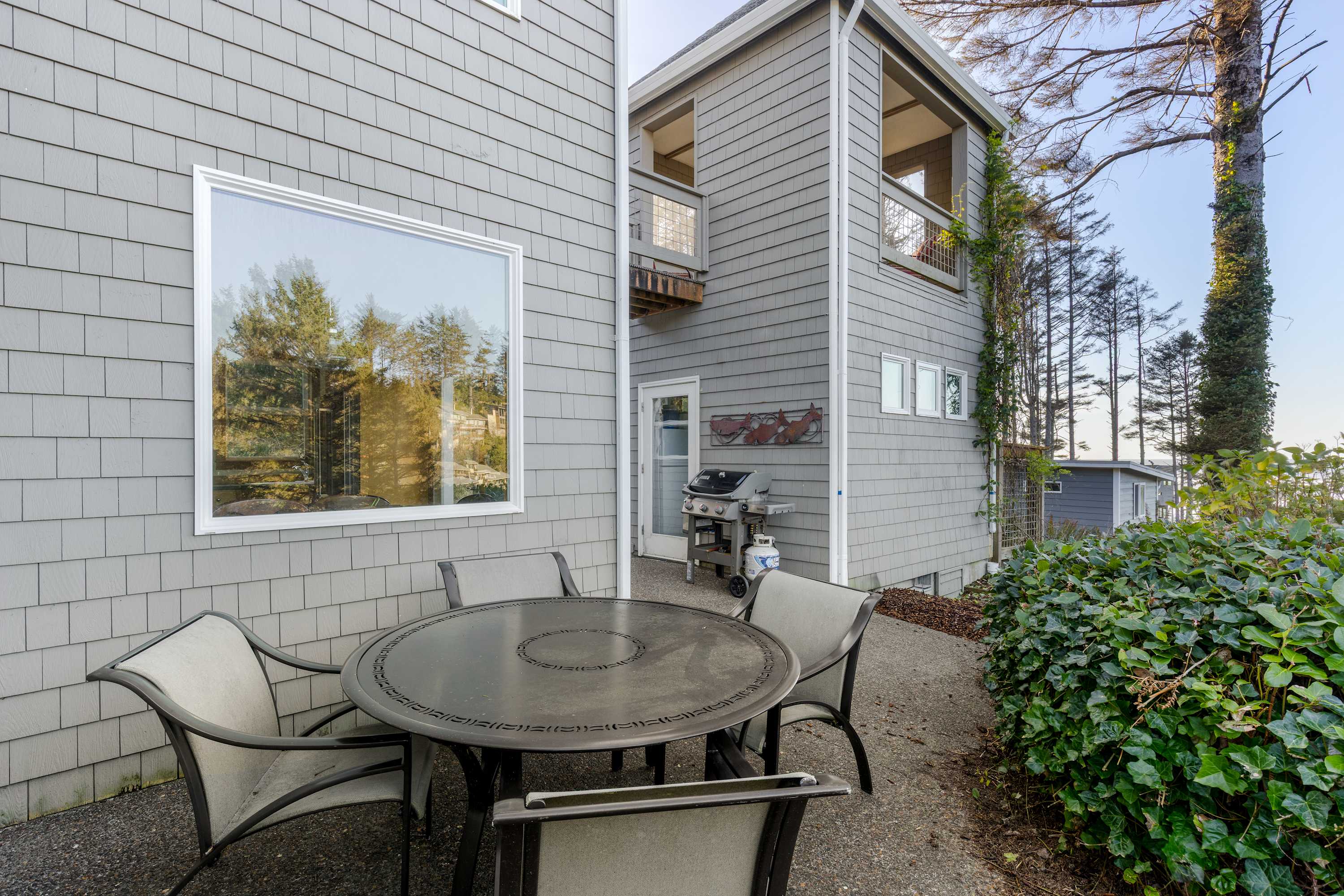 An outdoor patio area features a round metal dining table with four chairs on a concrete surface, adjacent to the gray shingled house. A gas grill is visible near a glass door, surrounded by trees and green foliage.