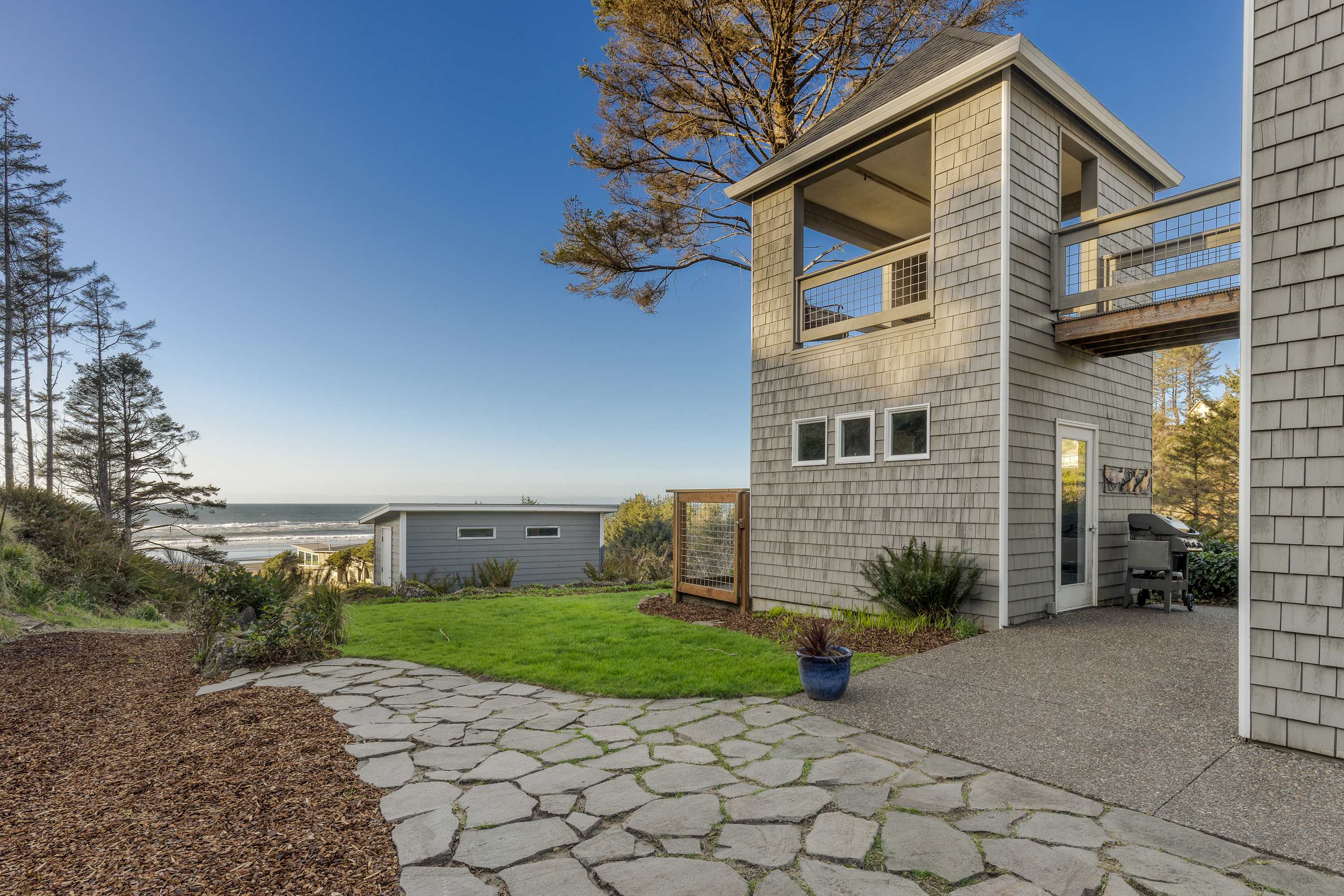 Rear view of the gray shingle-sided house, showcasing a multi-level structure with a lookout tower and an elevated walkway connecting to the main house. A stone path leads across a grassy lawn towards the ocean in the distance.