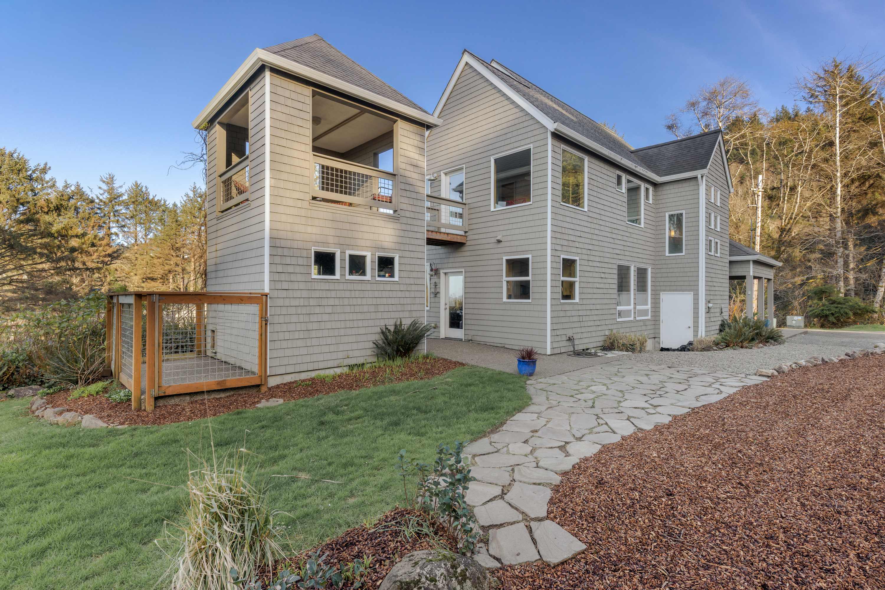 Rear view of the gray shingle-sided house, featuring multiple windows, a stone path, and a grassy lawn. A distinctive lookout tower with a balcony is visible on the left, and dense forest surrounds the property.