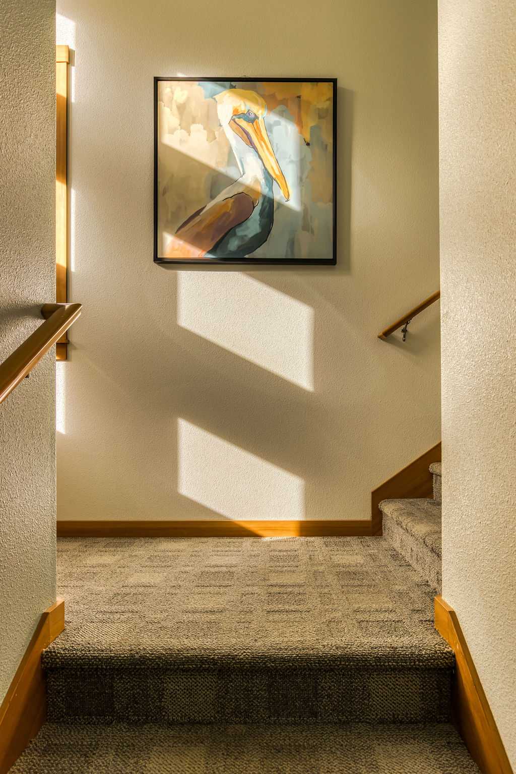 A carpeted staircase with wooden handrails leads to an upper level, illuminated by natural light from a nearby window. A large, colorful painting of a pelican serves as a focal point on the wall.
