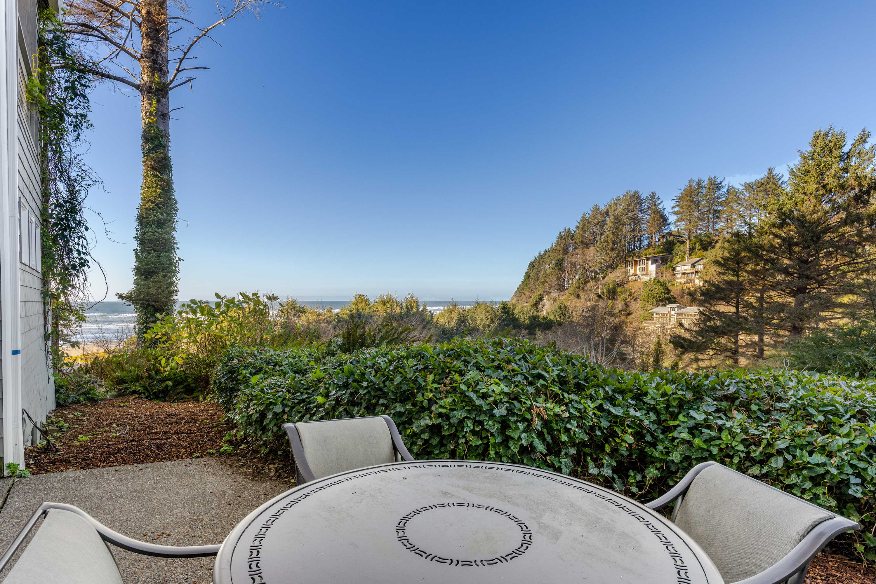 Outdoor patio area with a round metal table and three chairs, surrounded by lush green hedges and a tall tree. The ocean and a distant forested hillside are visible in the background, offering a scenic view.