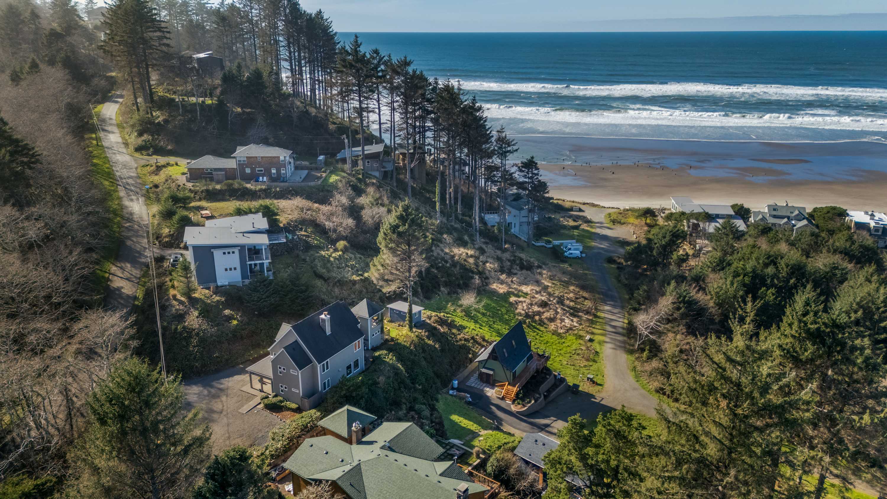 Aerial view showcasing the property and surrounding coastal landscape, with the gray house nestled among trees on a hillside. The Pacific Ocean and a sandy beach are prominent in the background, with other houses scattered along the coast.