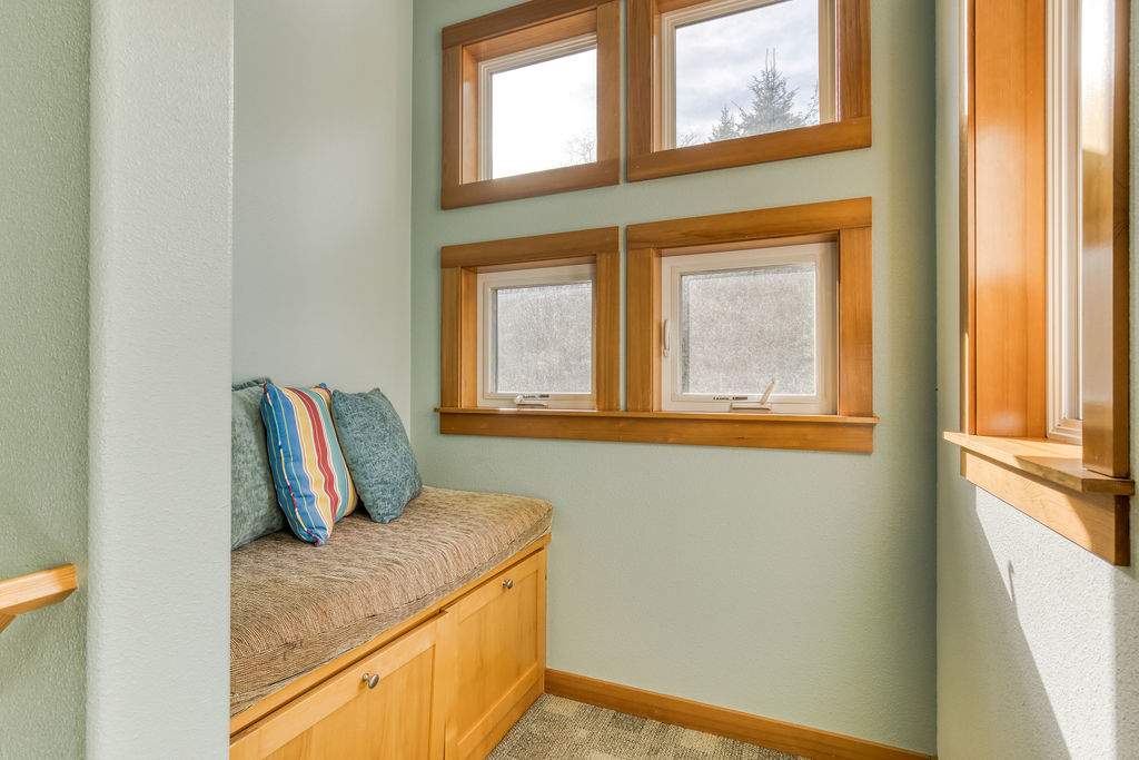 A cozy landing area features a built-in wooden bench with a comfortable cushioned seat and decorative throw pillows. Multiple windows with wooden frames allow ample natural light into the space, which has light blue-green walls.