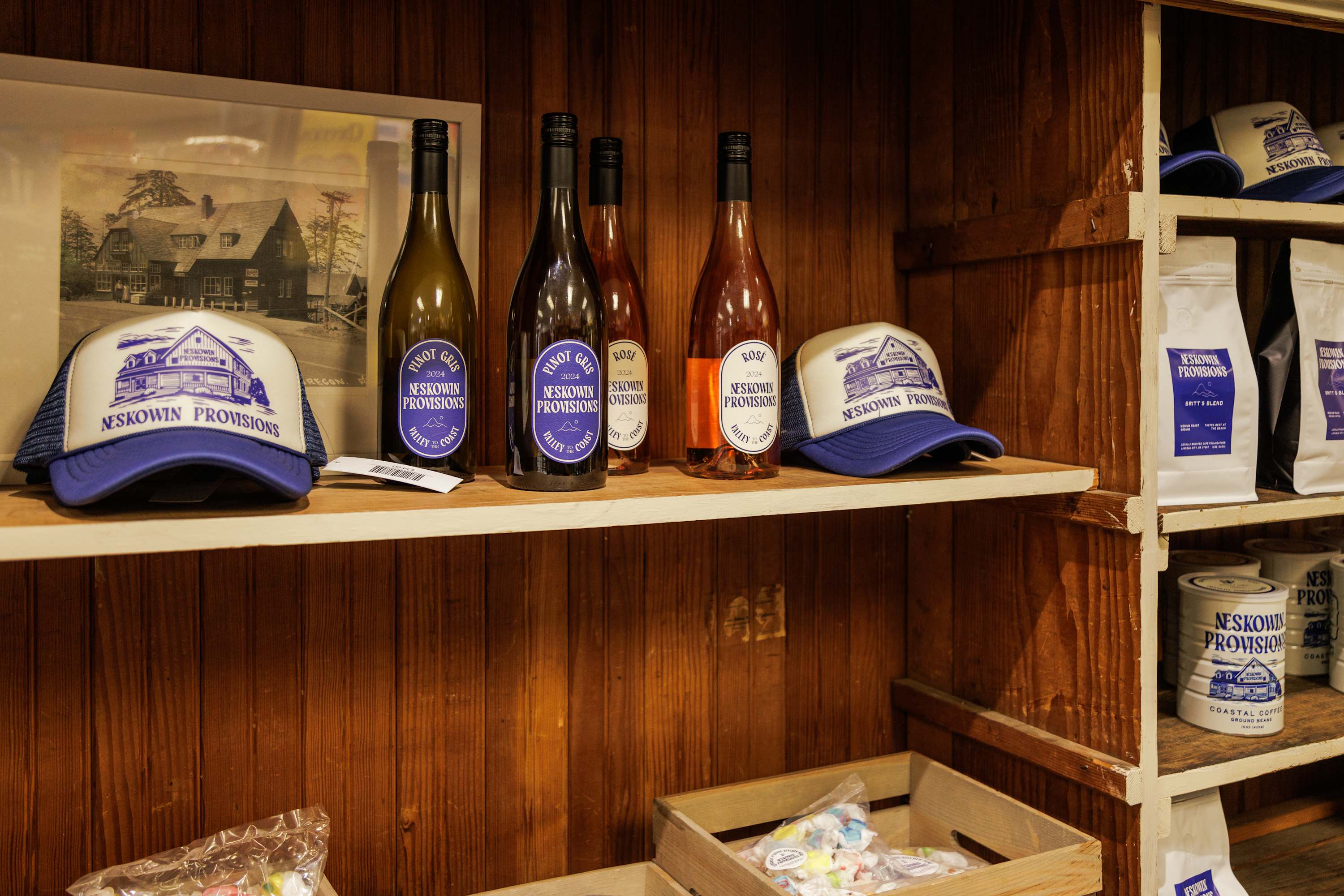 A rustic wooden shelf displaying bottles of wine, branded hats, and packaged coffee, all featuring the "NESKOWIN PROVISIONS" logo, suggesting a local shop or general store.
