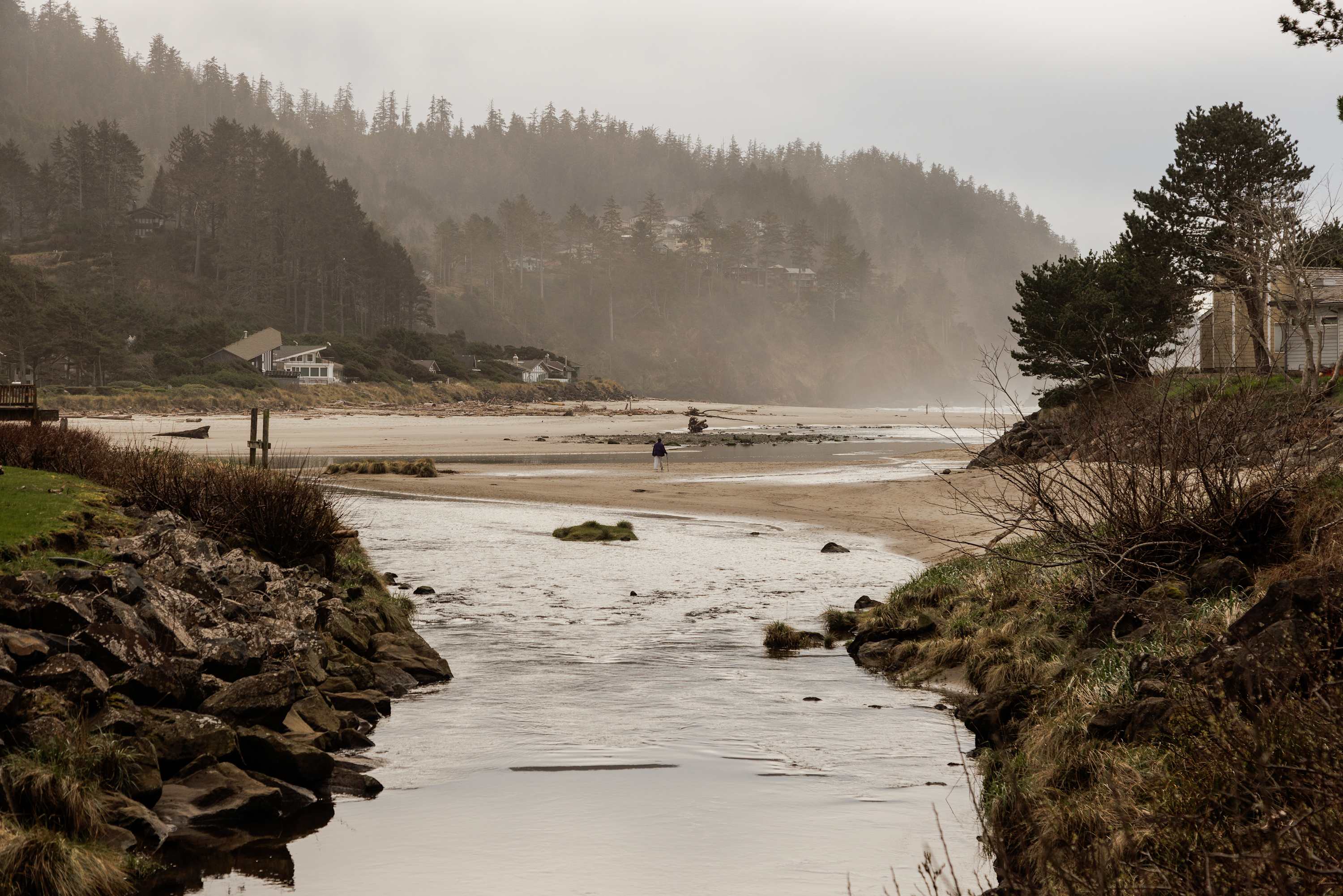 A misty, overcast view of a sandy beach with a narrow river flowing into the ocean, flanked by rocky banks and distant pine-covered hills with scattered houses. A lone person walks on the beach in the distance.