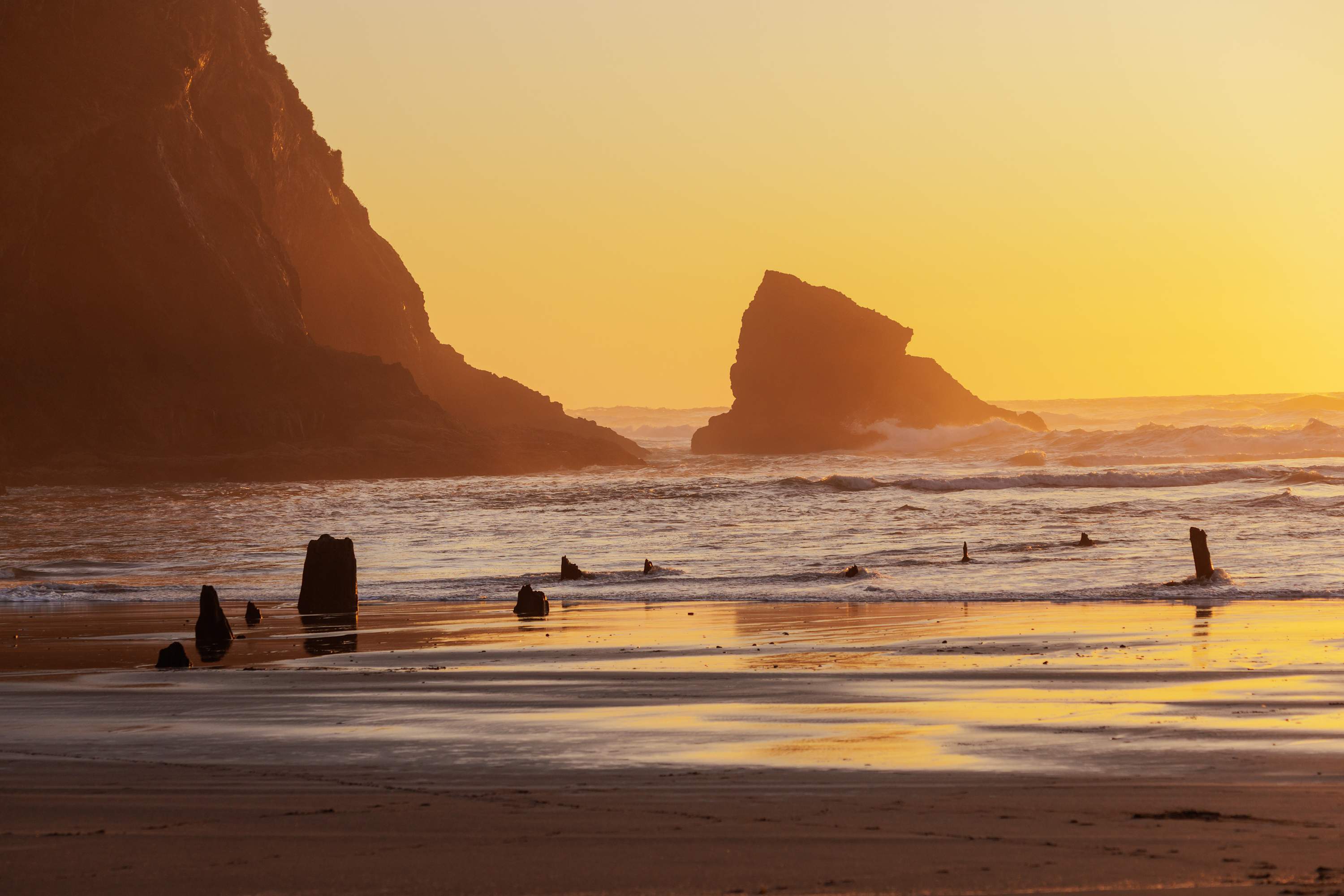 A serene sunset illuminates a sandy beach with large, dark rock formations silhouetted against a golden sky. Waves crash gently, reflecting the warm light, and several weathered wooden posts emerge from the water and sand.