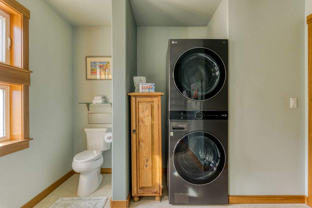 A section of bathroom-1 showing a white toilet, a small wooden cabinet, and a modern stacked washer and dryer unit, all against light blue walls.