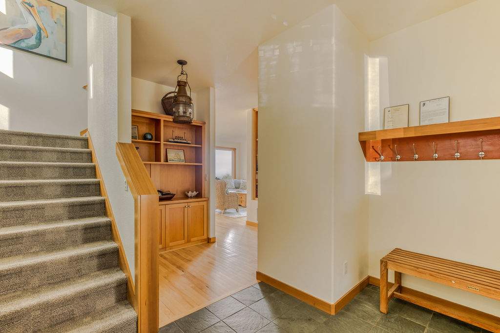 Bright foyer area with a carpeted staircase leading upstairs, featuring a wooden handrail. A built-in wooden bookshelf and a bench with coat hooks are visible, with a glimpse into another room with hardwood floors.