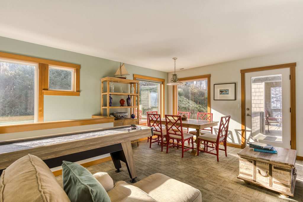 An inviting game room featuring a shuffleboard table, a light wood dining table with red chairs, and a comfortable sofa with throw pillows. Large windows with wooden trim offer views of the surrounding greenery, and a rustic wooden chest serves as a coffee table.