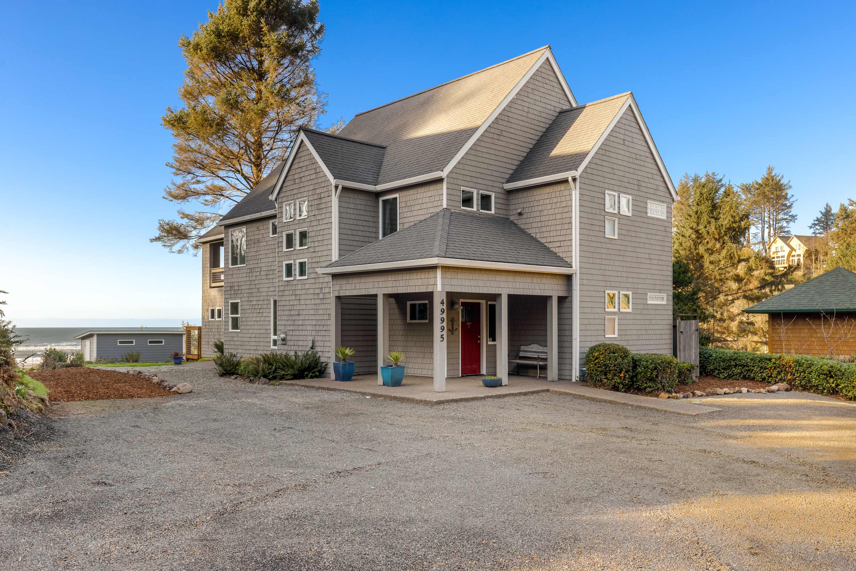 Front view of a large, multi-story gray shingle-sided house with a covered porch, red front door, and a gravel driveway. A tall evergreen tree stands to the left, and the ocean is visible in the distance.