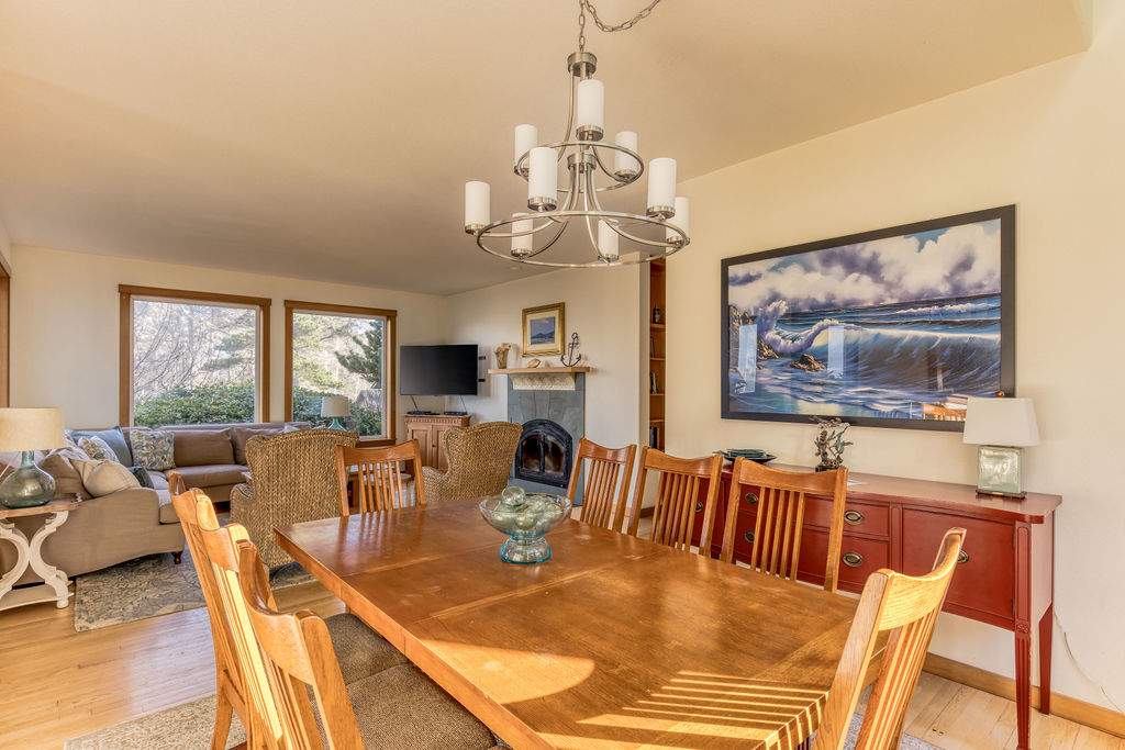 Spacious dining room with a long wooden table and eight chairs under a modern chandelier, with a large wave painting on the wall and the living room visible in the background through an open archway.