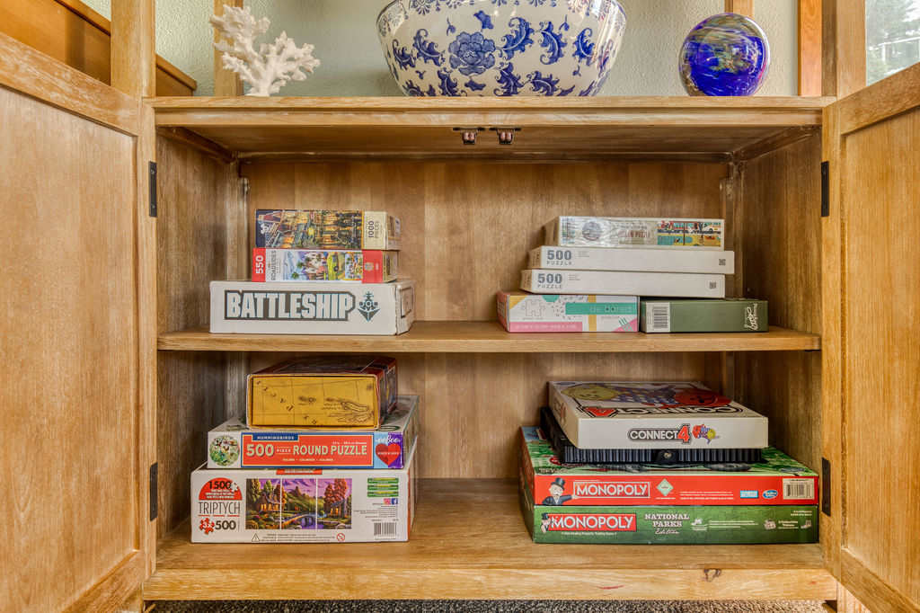A wooden cabinet filled with a variety of board games and puzzles, including Battleship, Monopoly, and Connect 4, neatly stacked on shelves. A large blue and white decorative bowl and a globe sit on top of the cabinet.