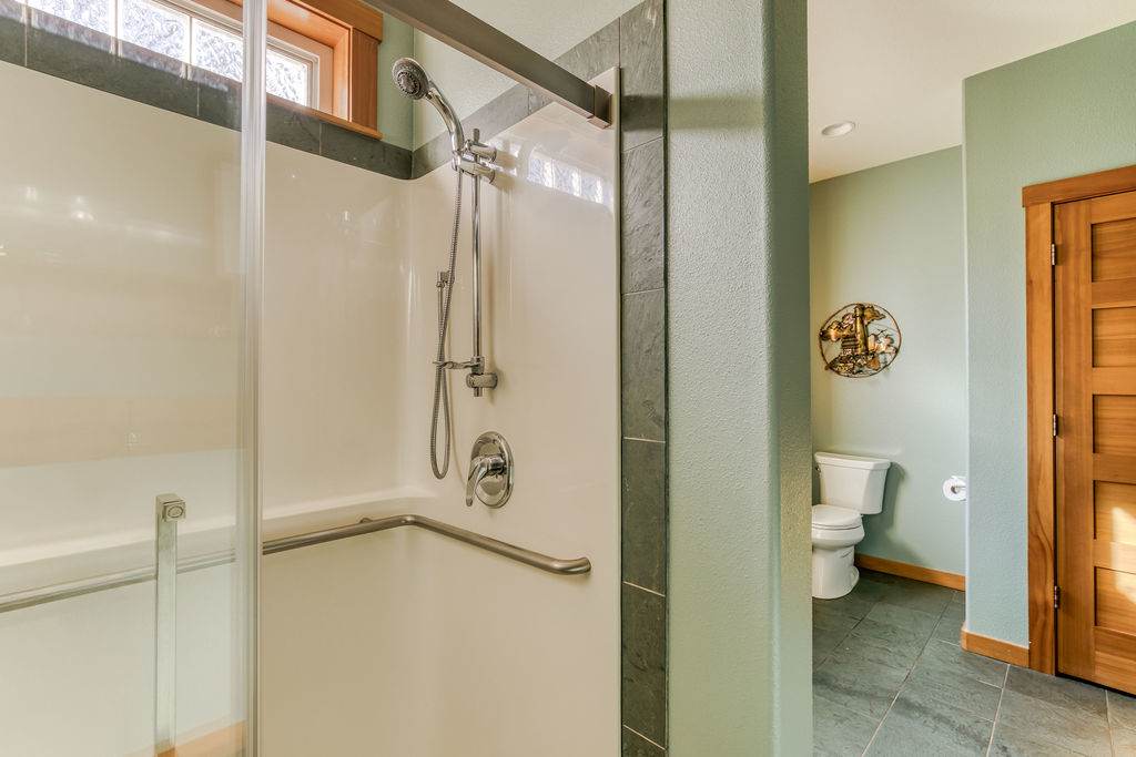 This view of the bathroom shows the toilet neatly tucked into an alcove, positioned next to the walk-in shower with its glass door and grab bar. The room maintains light green walls and dark tile flooring.
