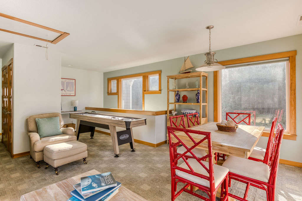 A spacious game room with a shuffleboard table, a light wood dining table with red chairs, and a comfortable armchair with an ottoman. Large windows with wooden frames overlook a green outdoor area, and a decorative shelf holds nautical-themed items.