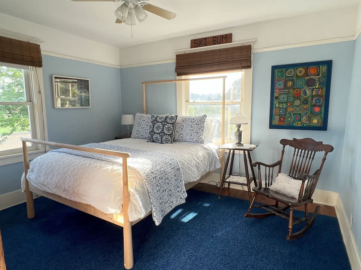 A cozy bedroom featuring light blue walls, a light wood bed frame with white and blue patterned bedding, and a dark blue carpet. A large window overlooks a bright outdoor scene, and a rocking chair sits beside a small table.