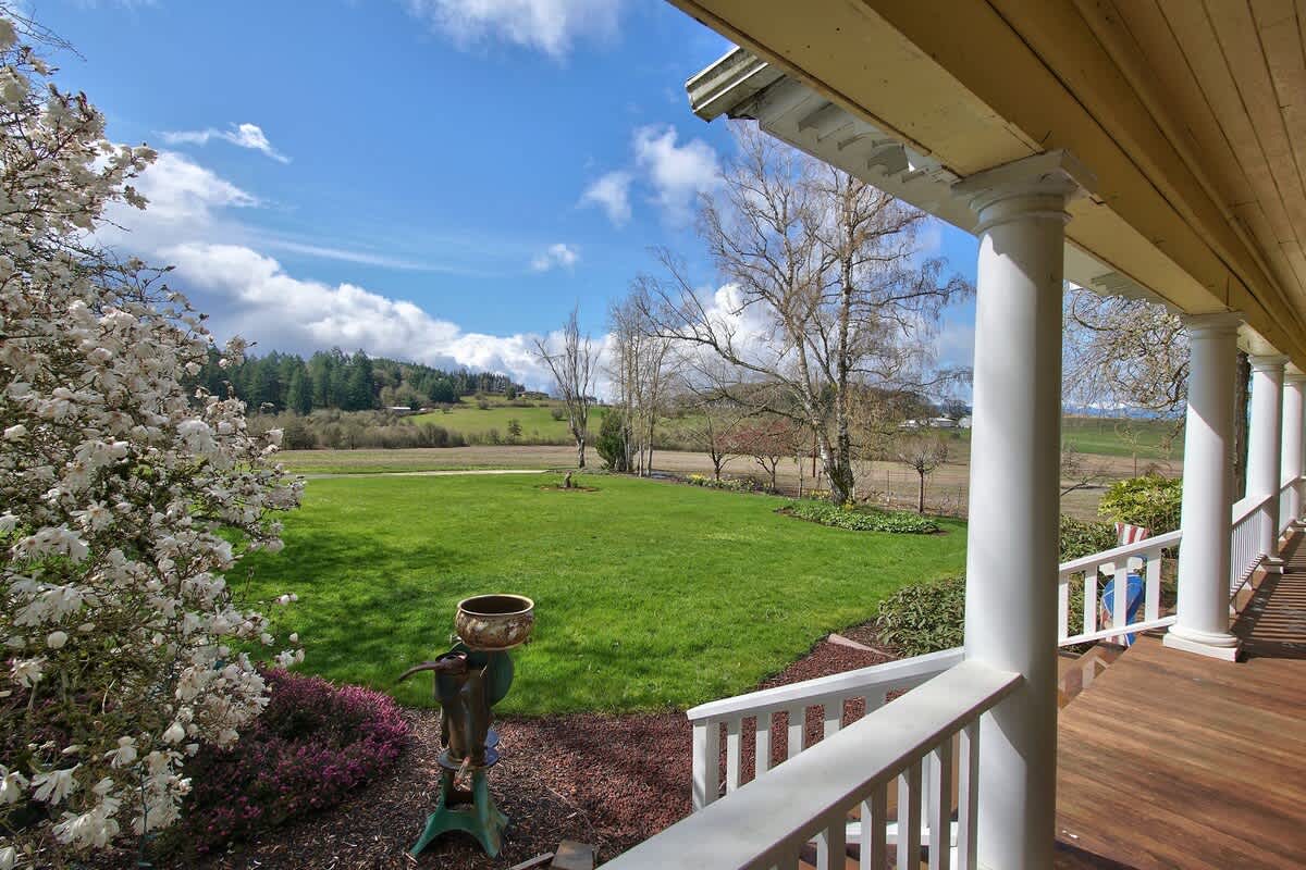 A scenic view from the porch overlooks a sprawling green lawn and distant rolling hills under a dynamic sky with white clouds, framed by flowering bushes and an antique water pump.