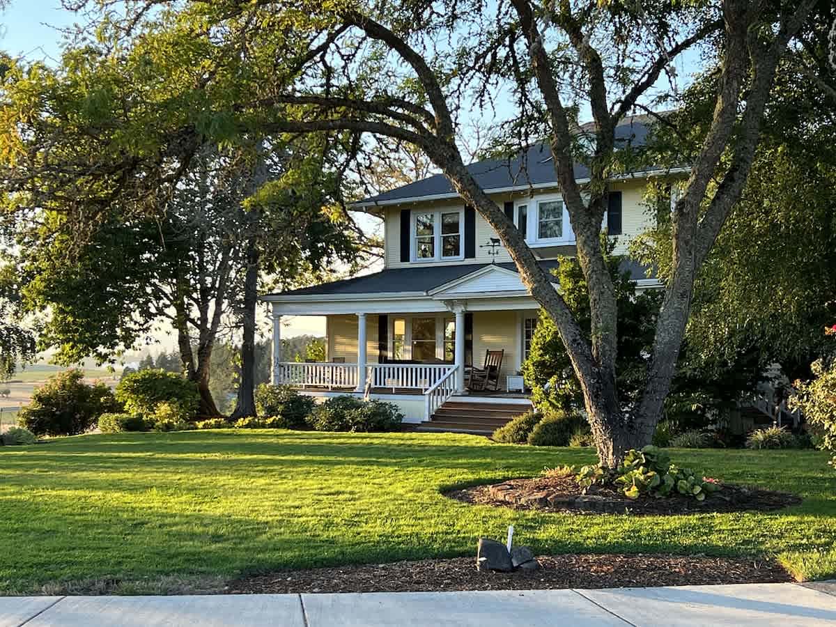A grand yellow two-story farmhouse with a spacious wrap-around porch and white railings, nestled among mature trees on a lush green lawn, bathed in warm evening sunlight.