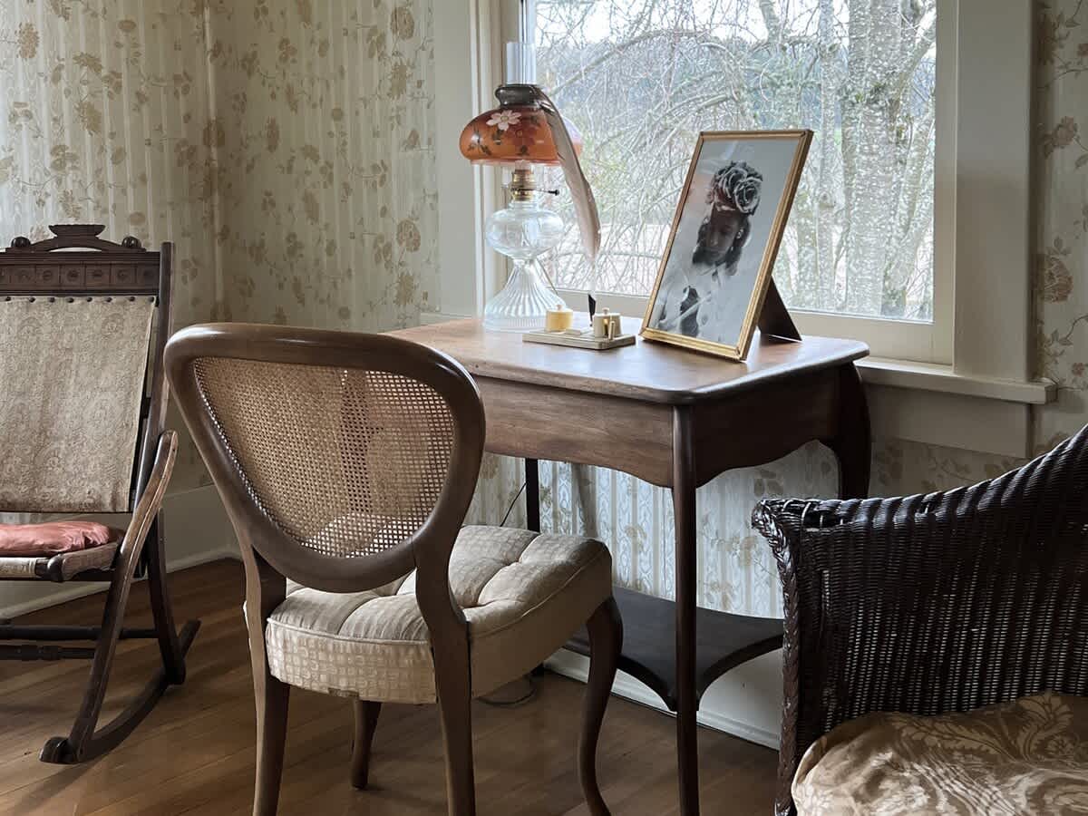 A cozy corner with a small wooden desk, a cane-back chair, and a vintage oil lamp, set against floral wallpaper with a window view of trees.