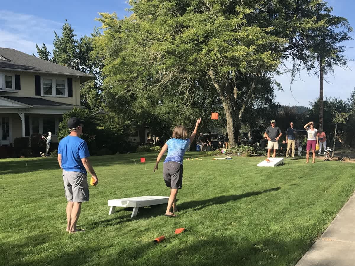 A vibrant green lawn with several people playing cornhole, surrounded by mature trees and the yellow farm home in the background, under a bright sunny sky.